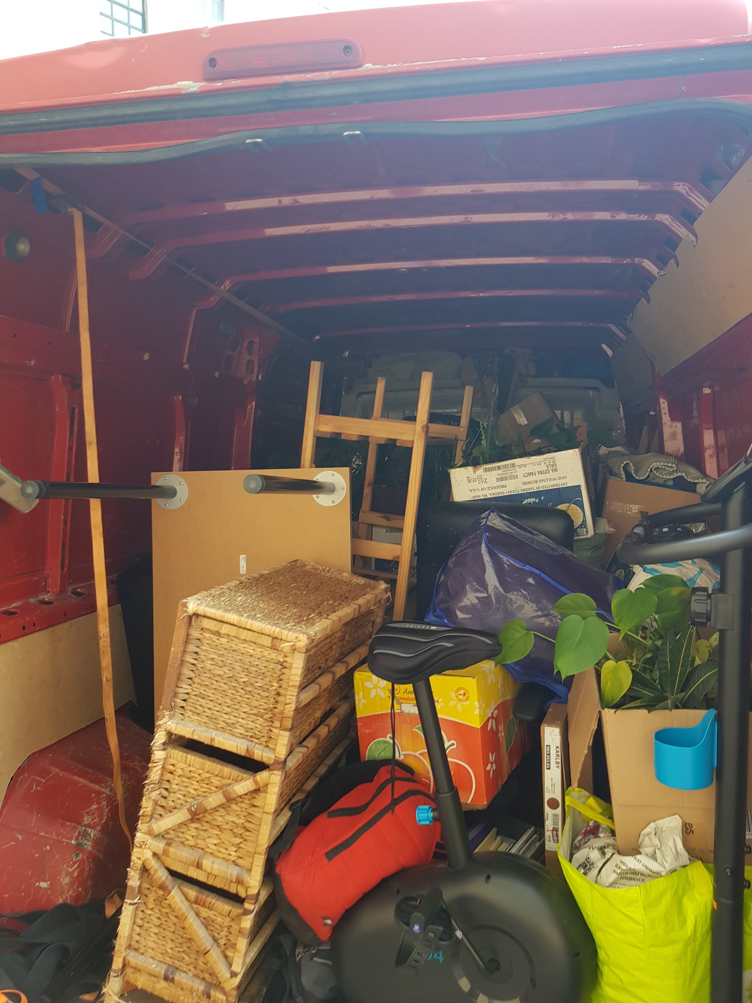 Wnętrze czerwonego busa wypełnione różnorodnymi przedmiotami podczas transportu, w tym wiklinowe kosze, drabina, stół z metalowymi nogami, rower treningowy, kartony, torba, roślina doniczkowa...