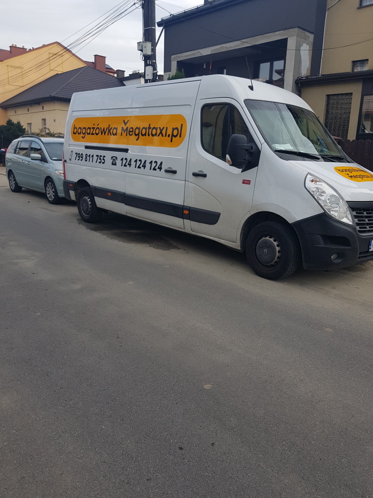 Biały bus dostawczy z żółtym logo 'bagażówka Megataxi.pl' i numerami telefonów zaparkowany na ulicy w pochmurny dzień.