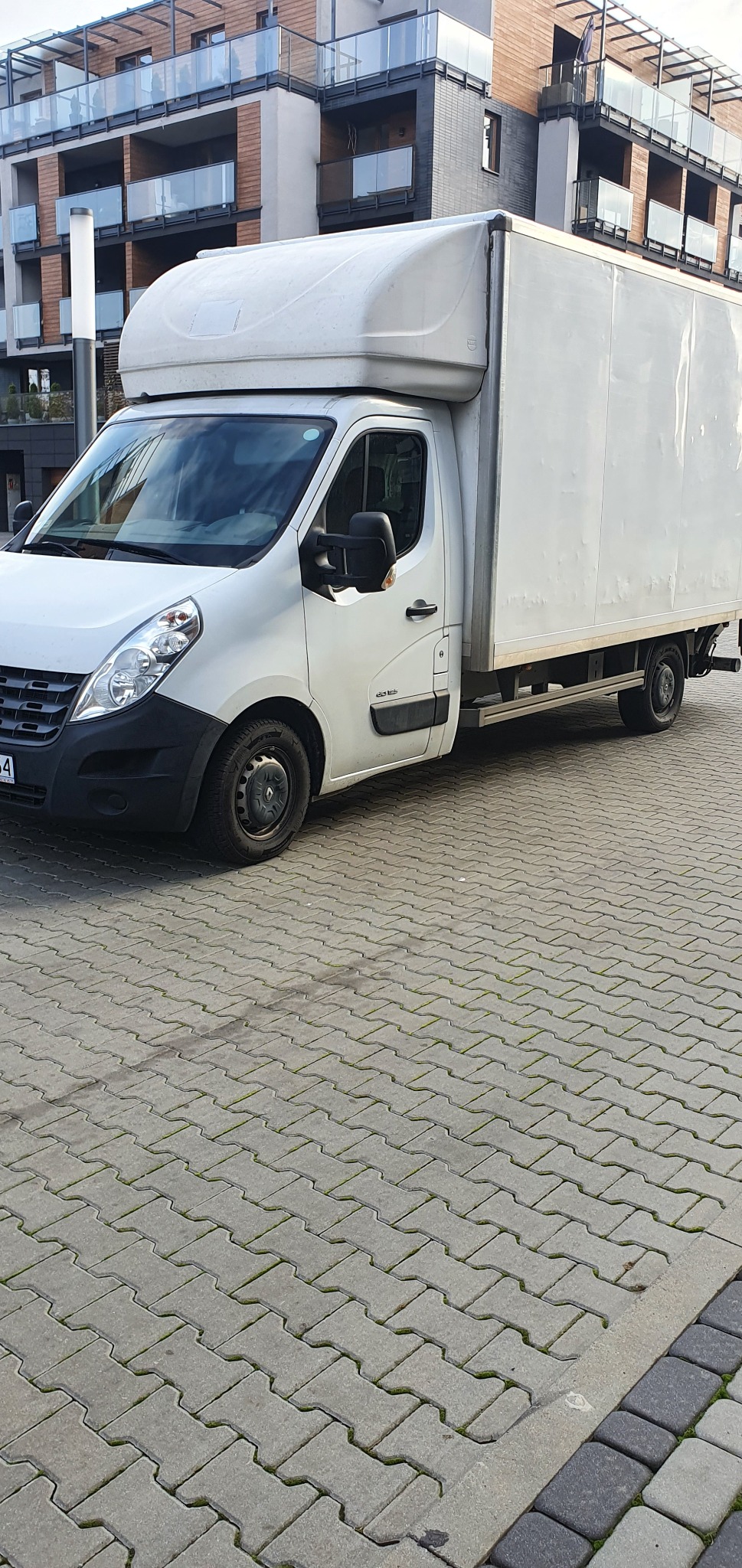 Biały samochód dostawczy Renault Master z wysoką zabudową na tle nowoczesnego budynku mieszkalnego, zaparkowany na szarej kostce brukowej.