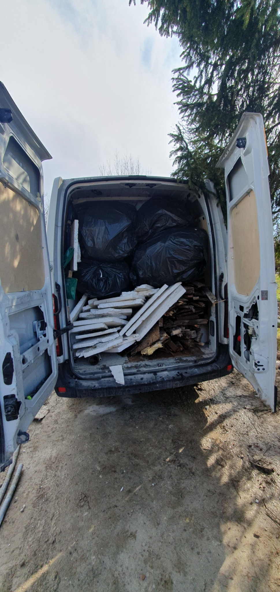 Otwarty bagażnik białego busa wypełniony czarnymi workami i stertą gruzu, w tle drzewo iglaste.