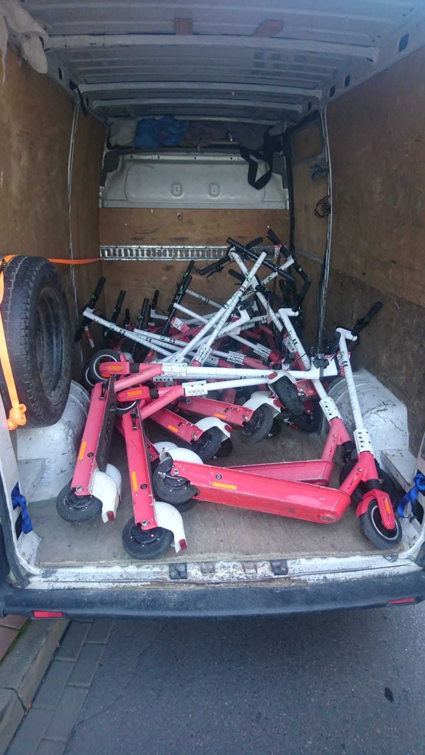 Wnętrze furgonetki wypełnionej stertą różowych i białych hulajnóg elektrycznych, z kołem zapasowym przymocowanym do boku, sugerujące transport lub relokację większej ilości pojazdów.