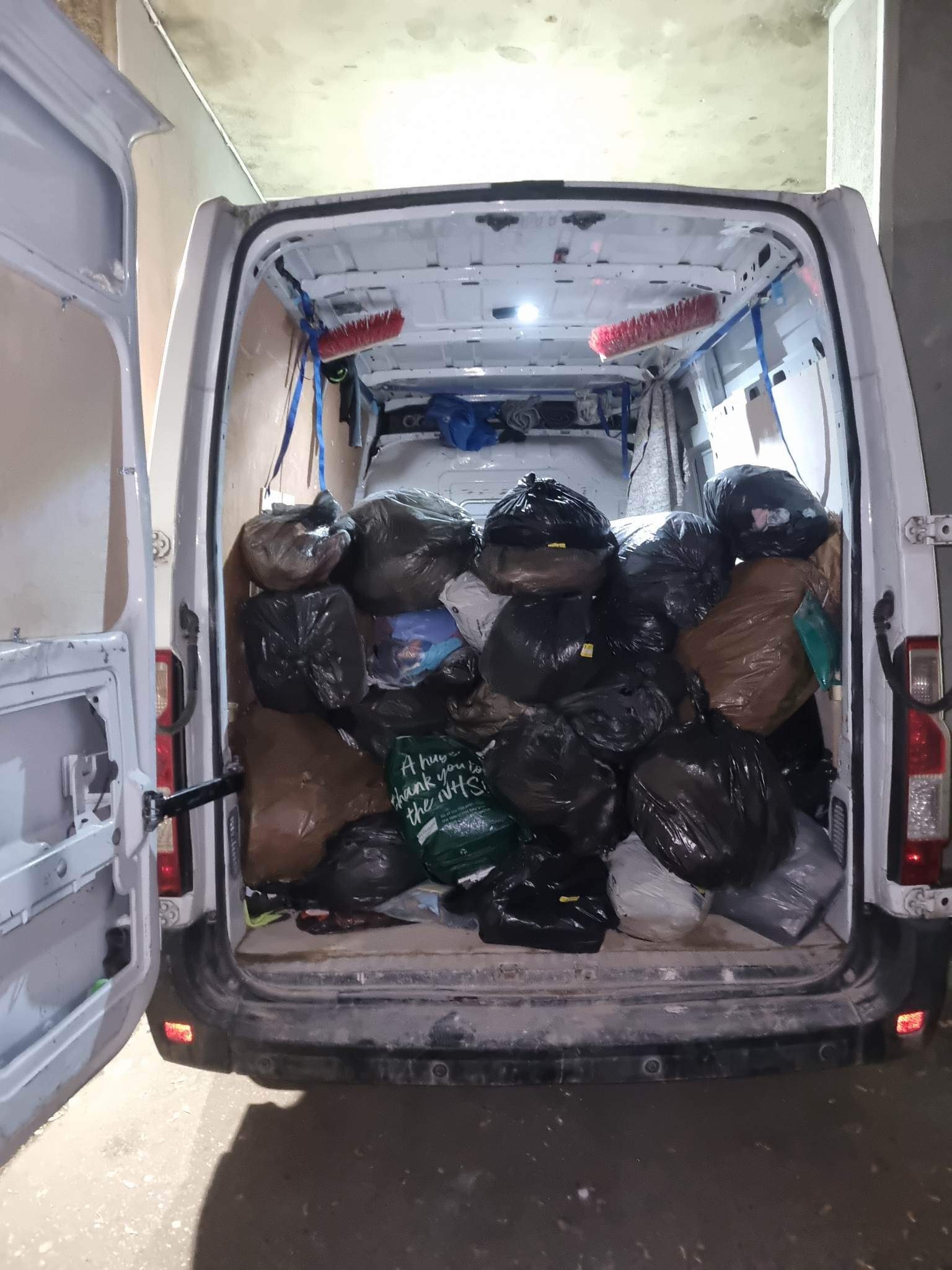 Wnętrze białego busa wypełnione workami z odpadami, widoczne dwie szczotki przymocowane do sufitu i torba z napisem 'A huge thank you to the NHS'.