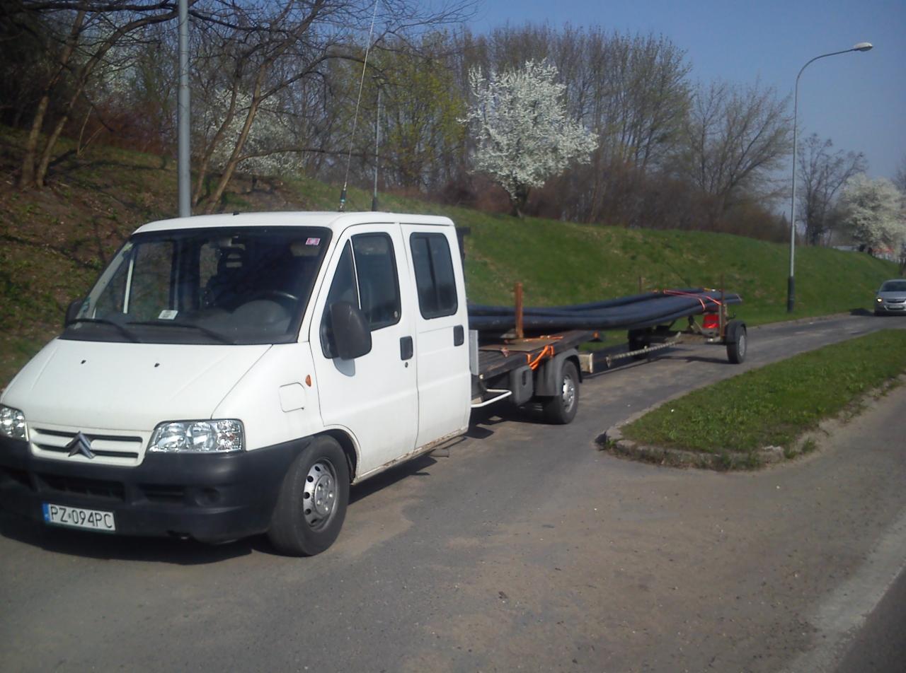 Biały samochód dostawczy Citroen z przyczepą przewożącą rury instalacyjne na tle wiosennego krajobrazu z kwitnącymi drzewami.