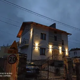 Dwupiętrowy dom z jasną elewacją oświetlony zewnętrznymi lampami ściennymi w kształcie wachlarza, widok na balkon z ozdobną balustradą, metalowa brama wjazdowa i znak drogowy z ograniczeniem...