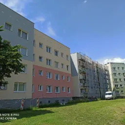 Wielorodzinny budynek z rusztowaniami, częściowo ocieplony i pomalowany na różne kolory, z zaparkowanym białym vanem na trawniku.