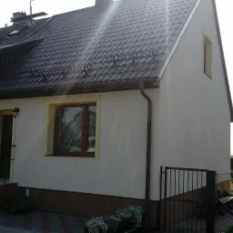 Dwukondygnacyjny dom po remoncie elewacji i wymianie dachu, z widocznymi elementami wykończeniowymi i nową dachówką w kolorze grafitowym.