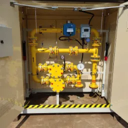 Żółta instalacja gazowa z rurami i zaworami, zamknięta w beżowej szafce z otwartymi drzwiami, widoczne liczniki i manometr, zabezpieczenie podłogi pasami ostrzegawczymi.