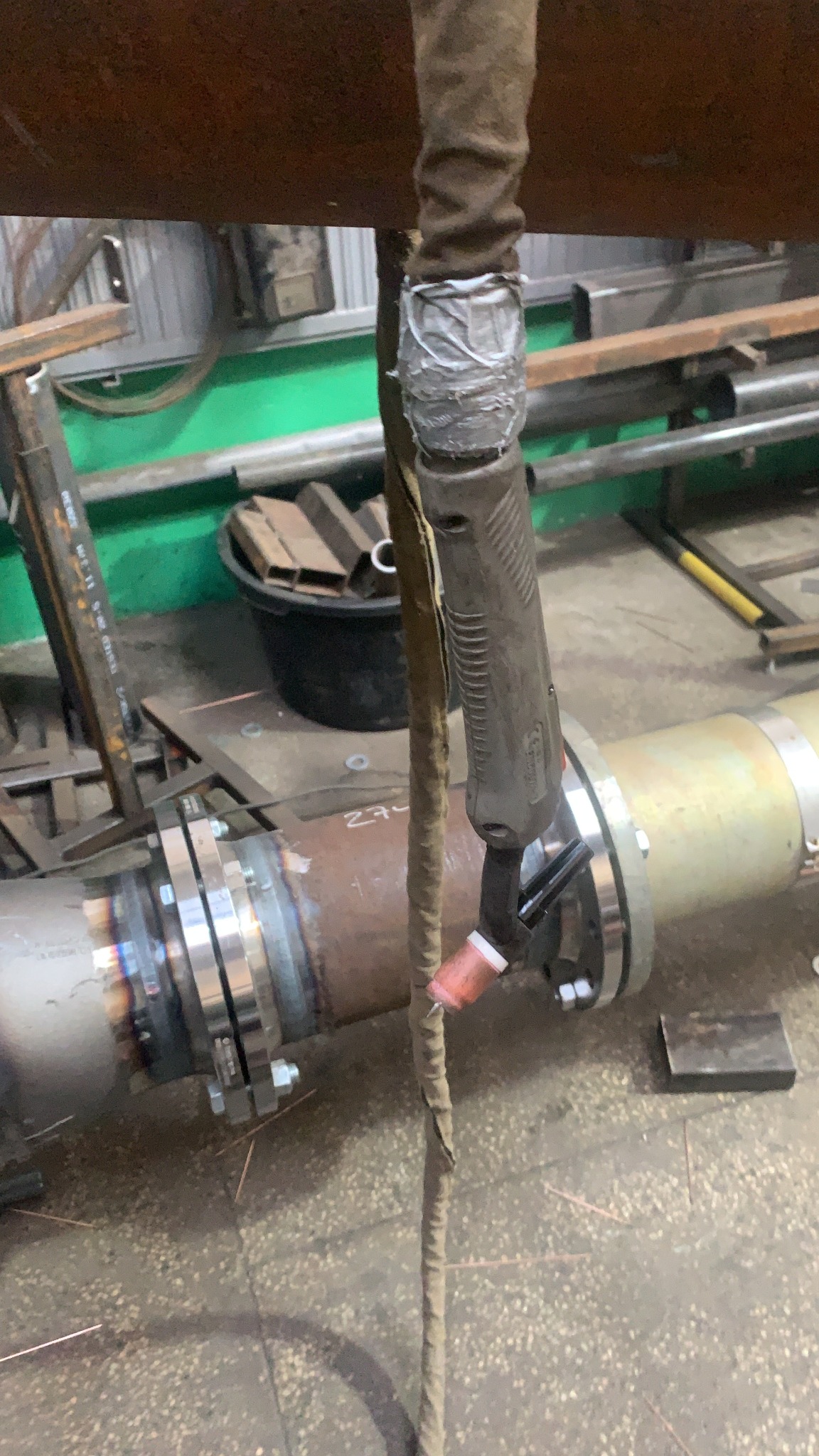 Przygotowanie do łączenia rur z kołnierzami metodą TIG; widoczny uchwyt spawalniczy z osłoną kabla owiniętą taśmą, spawana rura z widocznymi śladami ciepła i iskier na podłodze.