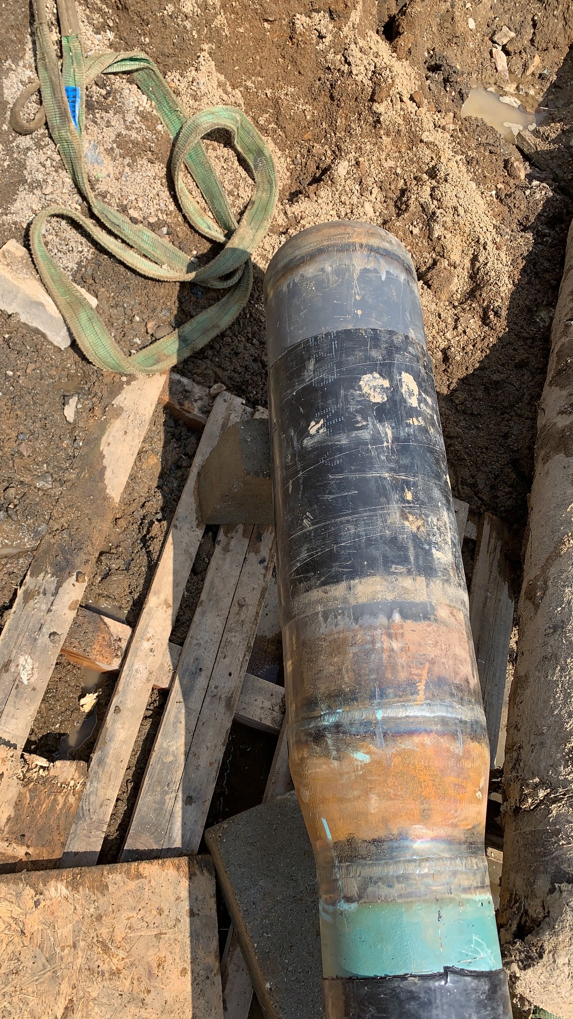 Rura z widocznymi śladami spawania i zieloną taśmą, leżąca na placu budowy obok drewnianych desek i zawiesia pasowego.