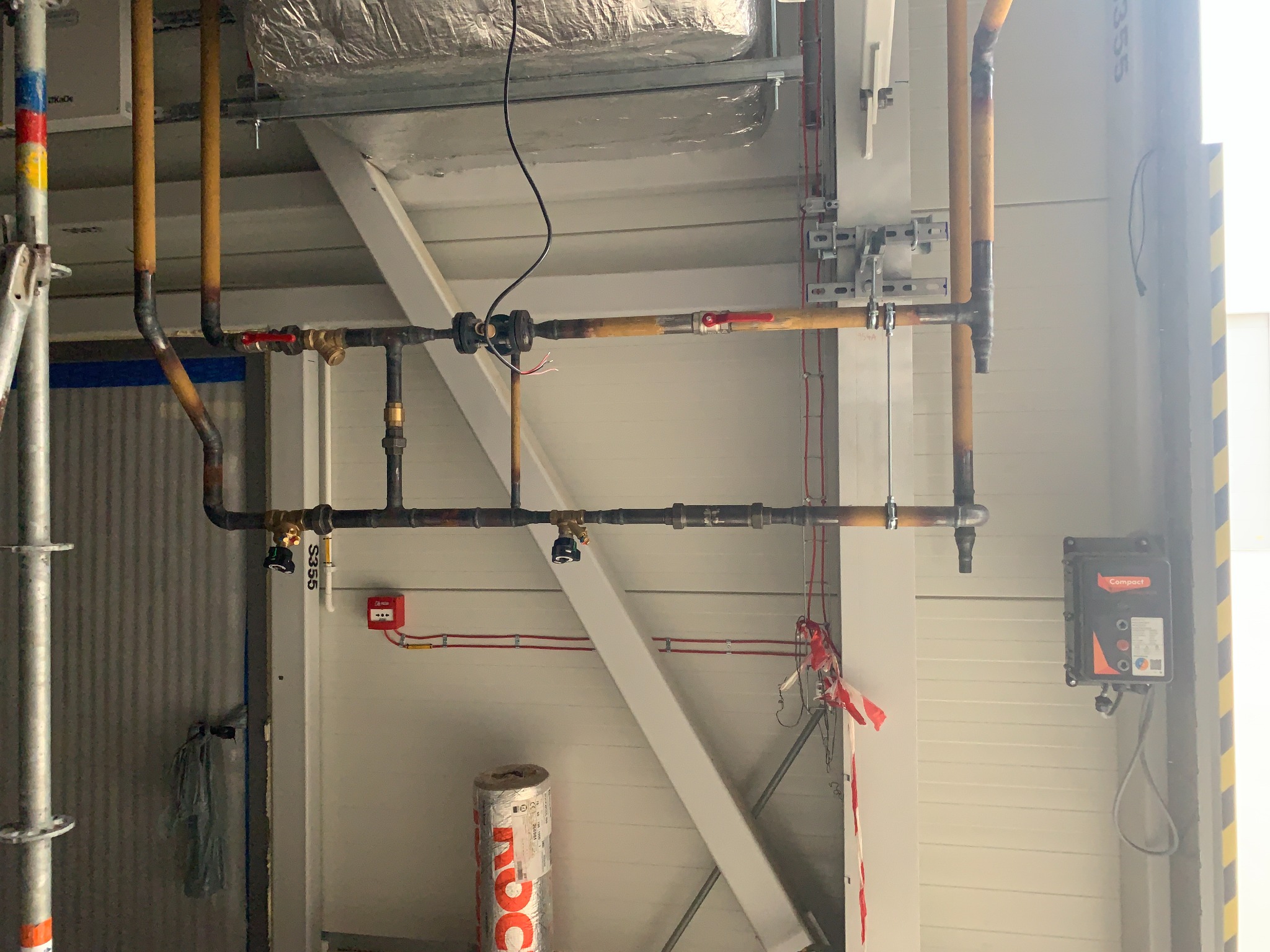 Instalacja rur miedzianych z zaworami, mocowana do sufitu w hali przemysłowej, widoczna instalacja elektryczna i element rusztowania.