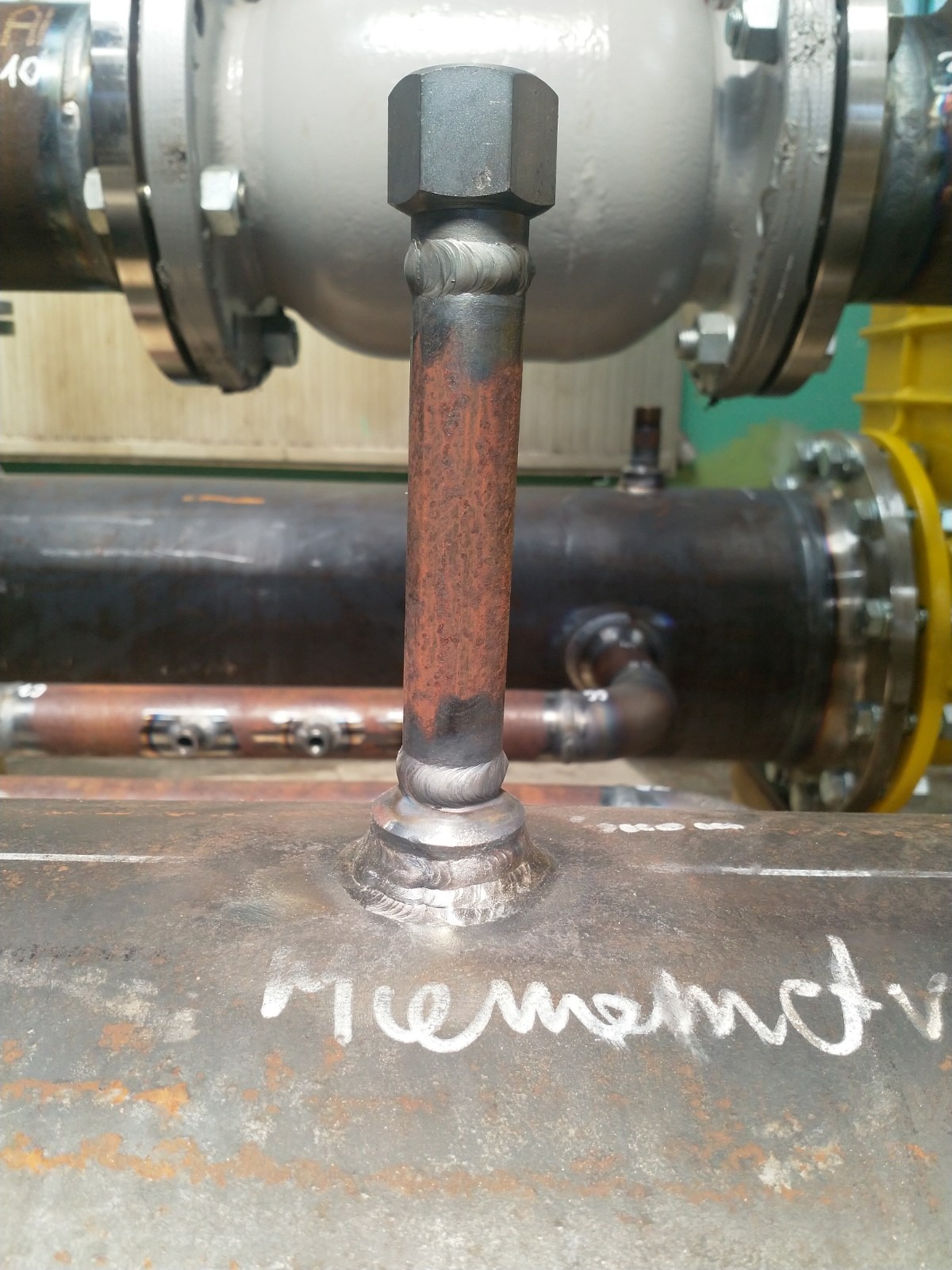 Spoina łącząca rdzewiejącą rurkę z grubą metalową rurą, z widocznymi śladami po spawaniu i napisem 'rememotr' na powierzchni rury, w tle elementy instalacji przemysłowej.