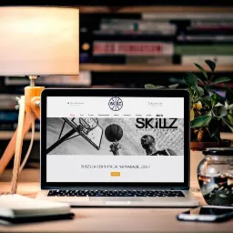 Laptop wyświetla stronę internetową z motywem koszykówki i hasłem 'Ruszyła rekrutacja na wakacje 2017!', stojący na drewnianym biurku obok drewnianej lampki, notatnika i rośliny doniczkowej,...