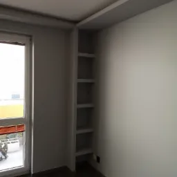Wnętrze pokoju po remoncie: białe ściany, ciemna podłoga, wbudowane półki obok okna z widokiem na balkon.