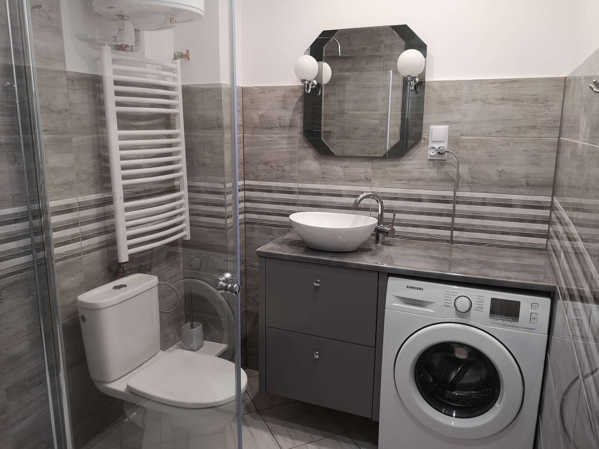Nowoczesna łazienka w szarościach z umywalką nablatową, pralką Samsung, grzejnikiem drabinkowym i ośmiokątnym lustrem z oświetleniem kulowym.