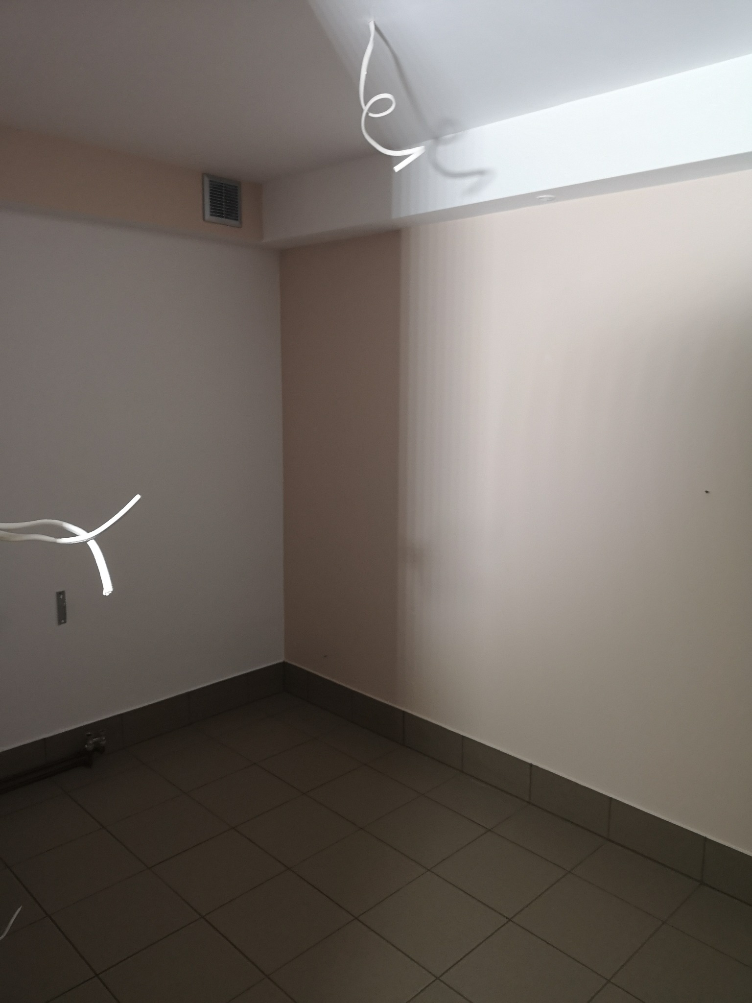 Puste pomieszczenie z szarą podłogą z płytek, beżowymi ścianami i wystającymi przewodami elektrycznymi z sufitu, widoczna kratka wentylacyjna.