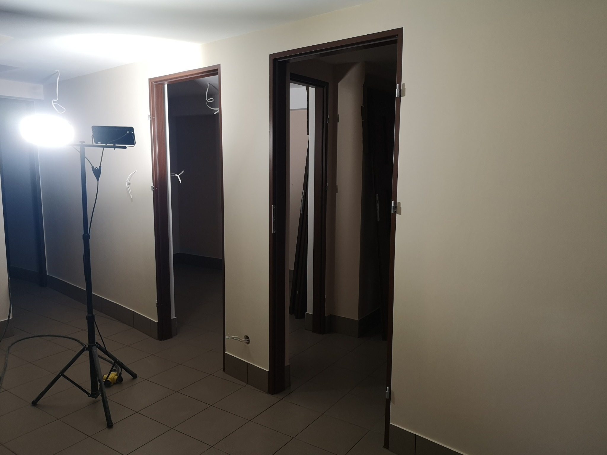 Wnętrze w trakcie remontu: widoczne trzy otwarte, ciemnobrązowe ościeżnice drzwiowe, szare płytki podłogowe, białe ściany, lampa budowlana na statywie oświetlająca korytarz, przewody elektryczne...
