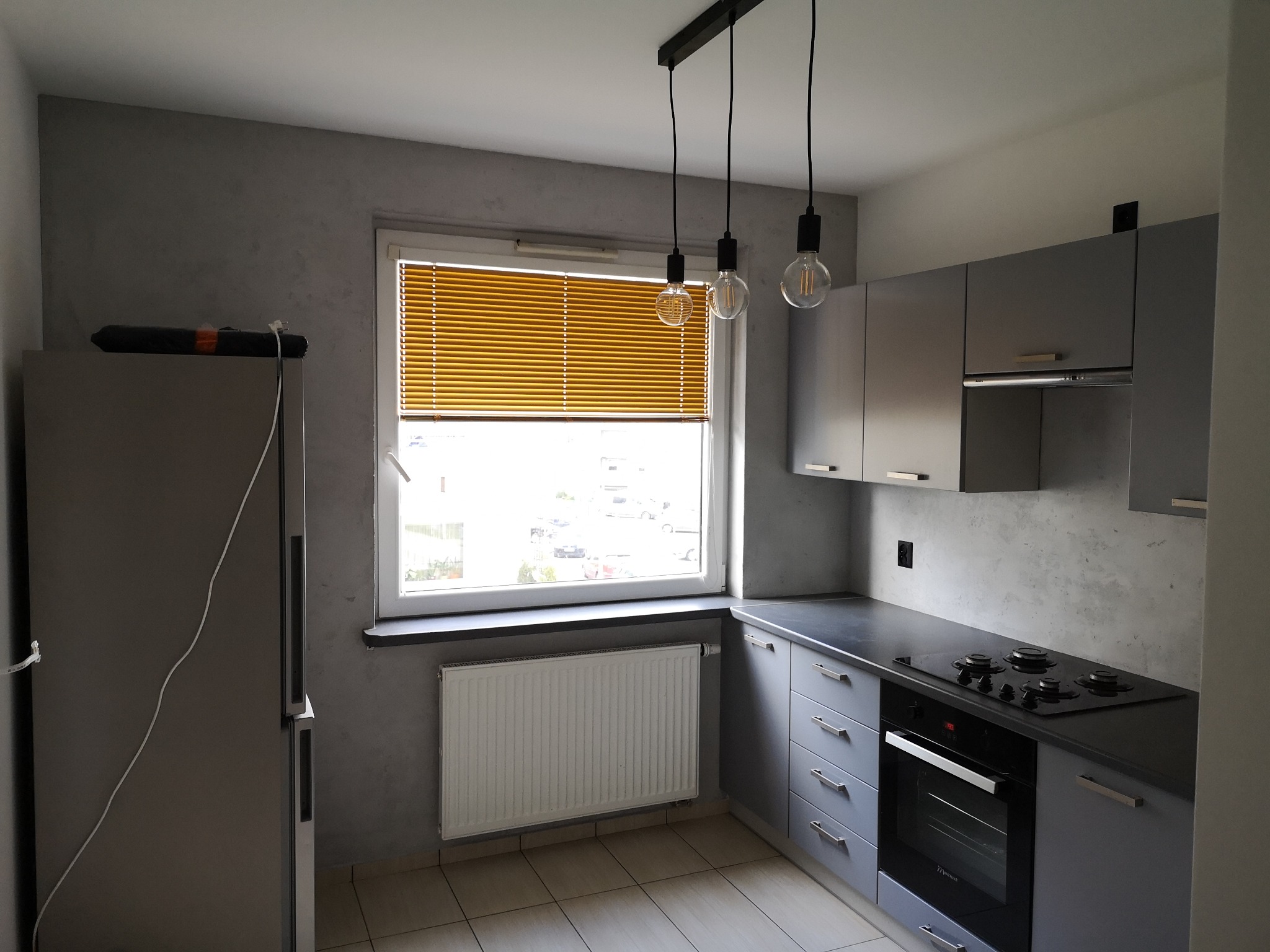 Nowoczesna kuchnia w odcieniach szarości z drewnianymi żaluzjami, wiszącymi lampami z żarówkami Edisona i płytą grzewczą.