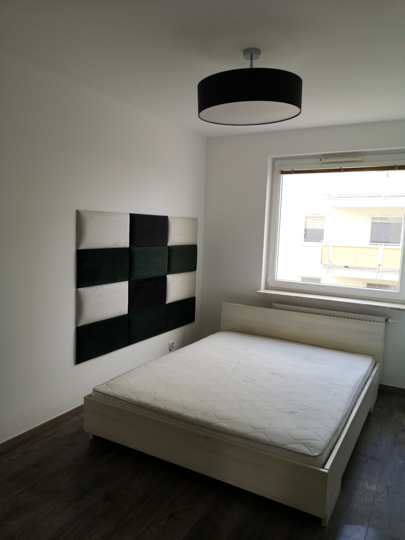 Proste łóżko z materacem w pokoju z panelami ściennymi w odcieniach bieli, szarości i zieleni, pod oknem i czarną lampą sufitową.