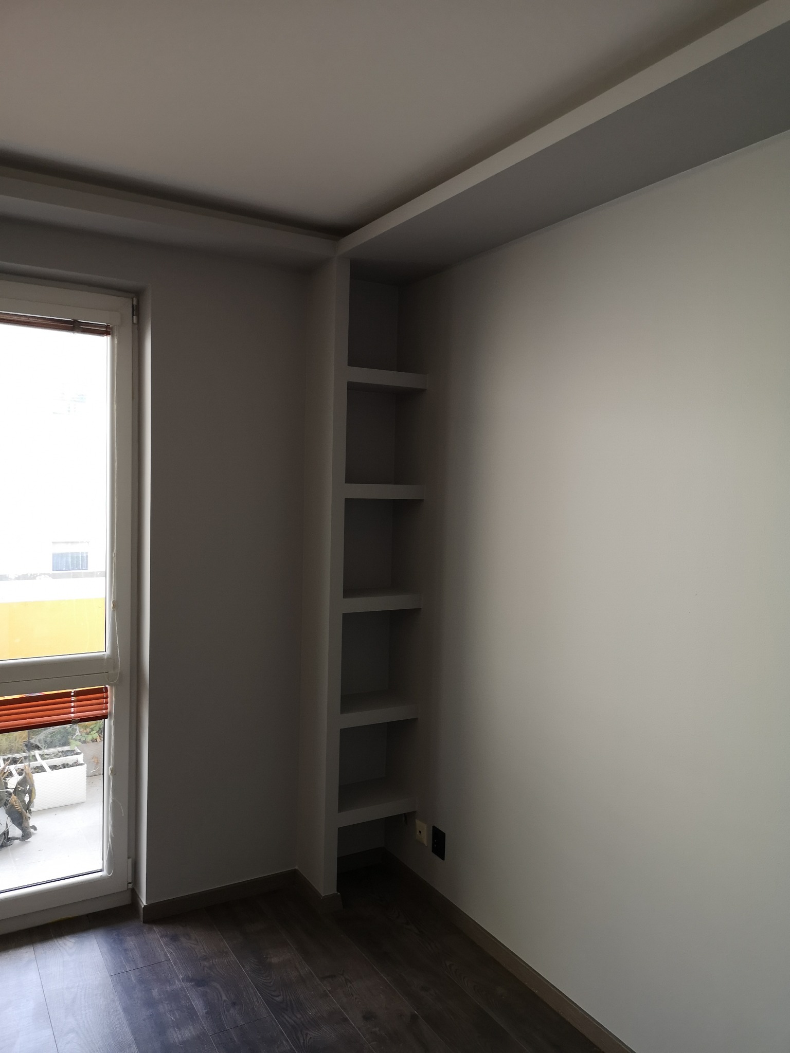 Wnętrze pokoju po remoncie: białe ściany, ciemna podłoga, wbudowane półki obok okna z widokiem na balkon.