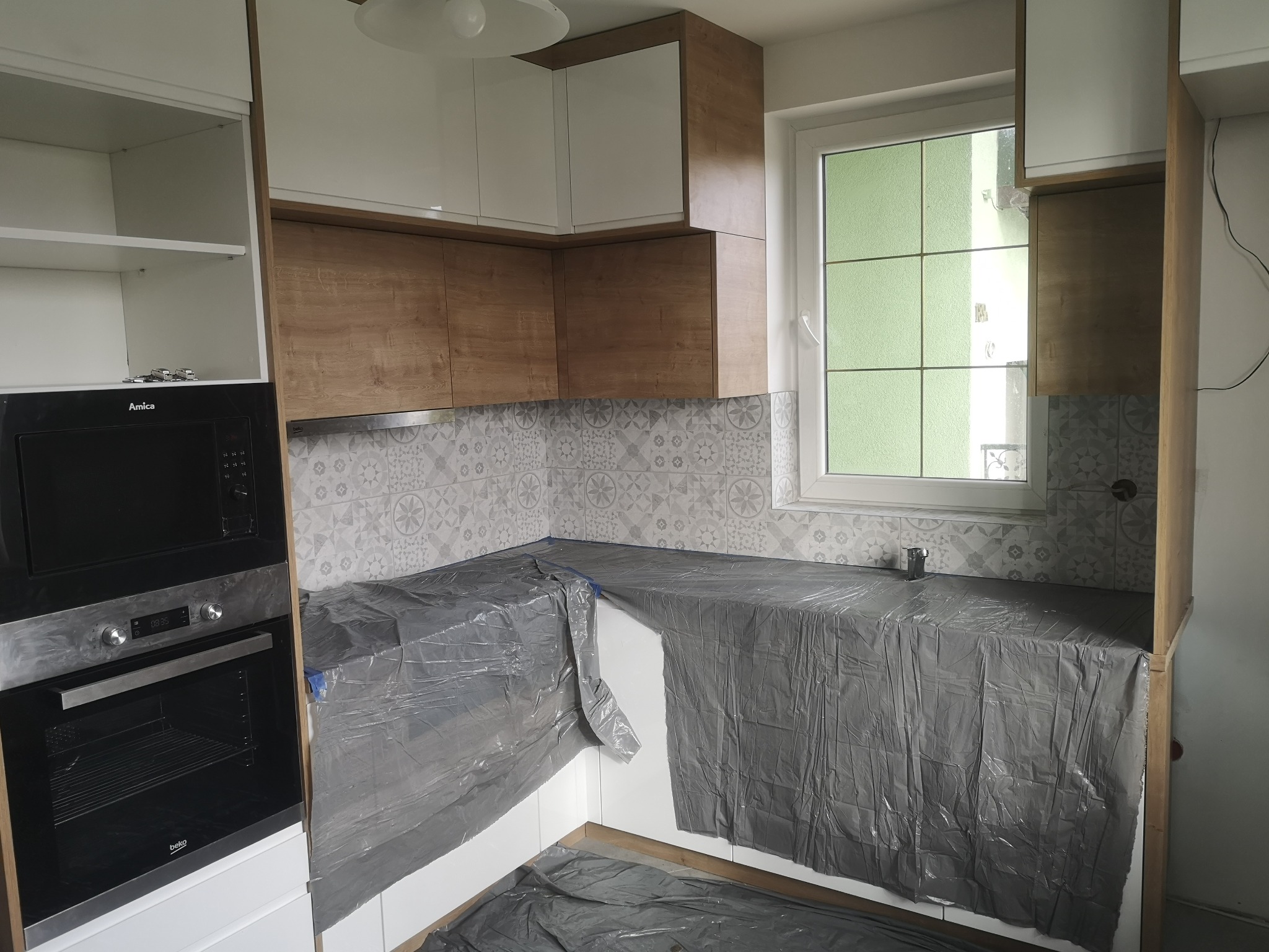 Kuchnia w trakcie zabezpieczania folią malarską przed pracami wykończeniowymi, widoczne zabudowane szafki górne białe i dolne w kolorze drewna, płytki z wzorem na ścianie, okno z widokiem na zieleń.