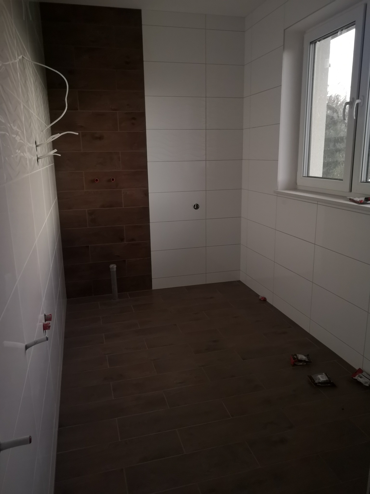 Wnętrze łazienki w trakcie wykańczania, z częściowo ułożonymi białymi i brązowymi płytkami, widoczne wystające rury i kable, okno z widokiem na zewnątrz oraz resztki opakowań po materiałach...