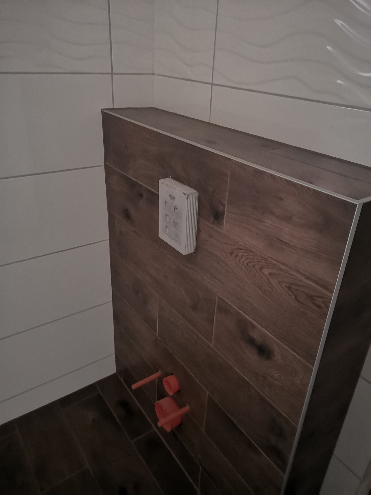 Stelaż podtynkowy WC obudowany płytkami imitującymi drewno, widoczne przyłącza wodne i przycisk spłukujący Grohe, fragment ściany wyłożonej białymi płytkami.