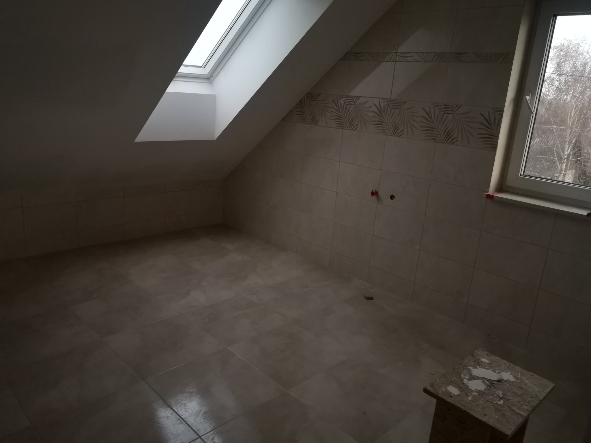 Ukończona łazienka na poddaszu w Bohutynie, wykończona beżowymi płytkami z motywem liści, widoczne okno dachowe i standardowe okno, na podłodze płytki w tym samym odcieniu, w rogu stoi mały stołek...