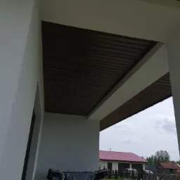 Zadaszenie ganku z brązową podbitką i białymi filarami, widok na dom z czerwonym dachem i rozwieszone pranie.