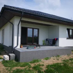 Nowoczesny dom murowany z ciemną dachówką i szarą elewacją tarasu, na którym stoi mały pies rasy sznaucer miniaturowy obok skrzynki z sadzonkami.