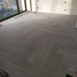 Ułożona pod kątem jasna podłoga z płytek imitujących drewno w surowym wnętrzu z dużym oknem i ścianą przygotowaną do malowania.