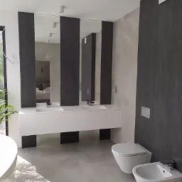 Nowoczesna łazienka z podwójną umywalką na białej szafce, wiszącą toaletą i bidetem, wykończona płytkami w odcieniach szarości i beżu, z dużym oknem i odbiciem sypialni w lustrze.