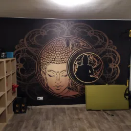 Ściana z fototapetą przedstawiającą wizerunek Buddy w kolorach złota i czerni, obok regał z książkami i sprzęt do ćwiczeń.