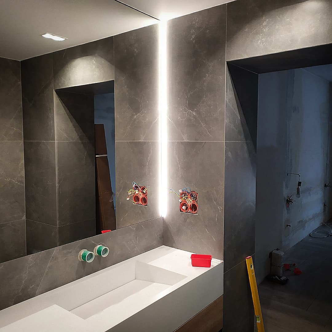 Łazienka w trakcie remontu z szarymi płytkami imitującymi marmur, podświetlane lustro z widocznymi instalacjami elektrycznymi, biała umywalka i poziomica oparta o ścianę.