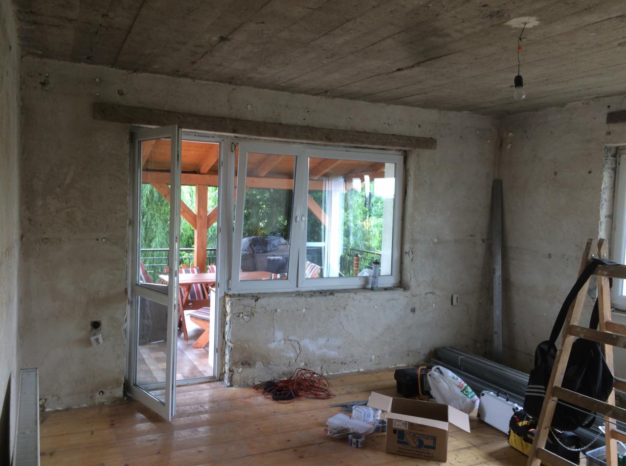 Surowy pokój w trakcie remontu z otwartym oknem balkonowym na zewnątrz, widoczne narzędzia i materiały budowlane na drewnianej podłodze, ściany w stanie surowym przed wykończeniem.