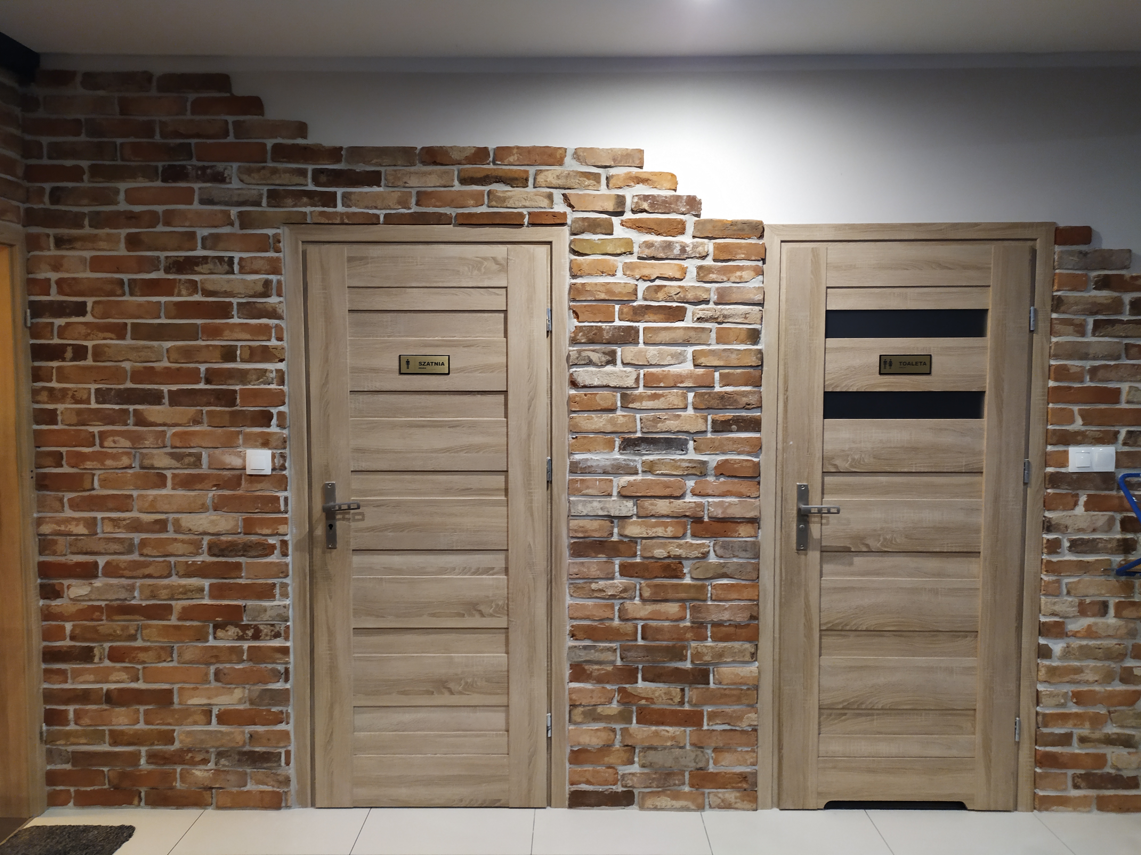 Wykończenie ściany z cegły klinkierowej wokół dwóch drewnianych drzwi z tabliczkami: szatnia i toaleta. Cegła ułożona do pewnej wysokości ściany, tworząc nieregularne zakończenie.