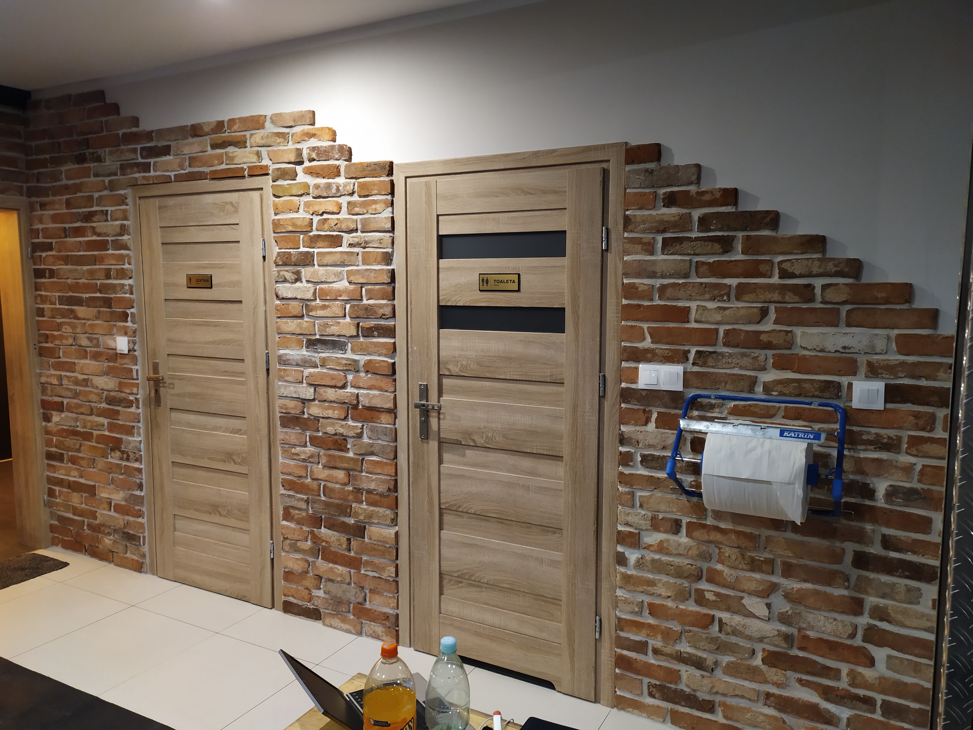 Dwie drewniane drzwi z oznaczeniami toalet (damska i męska) na ścianie wyłożonej cegłą w stylu rustykalnym, z fragmentem laptopa i butelkami na pierwszym planie.