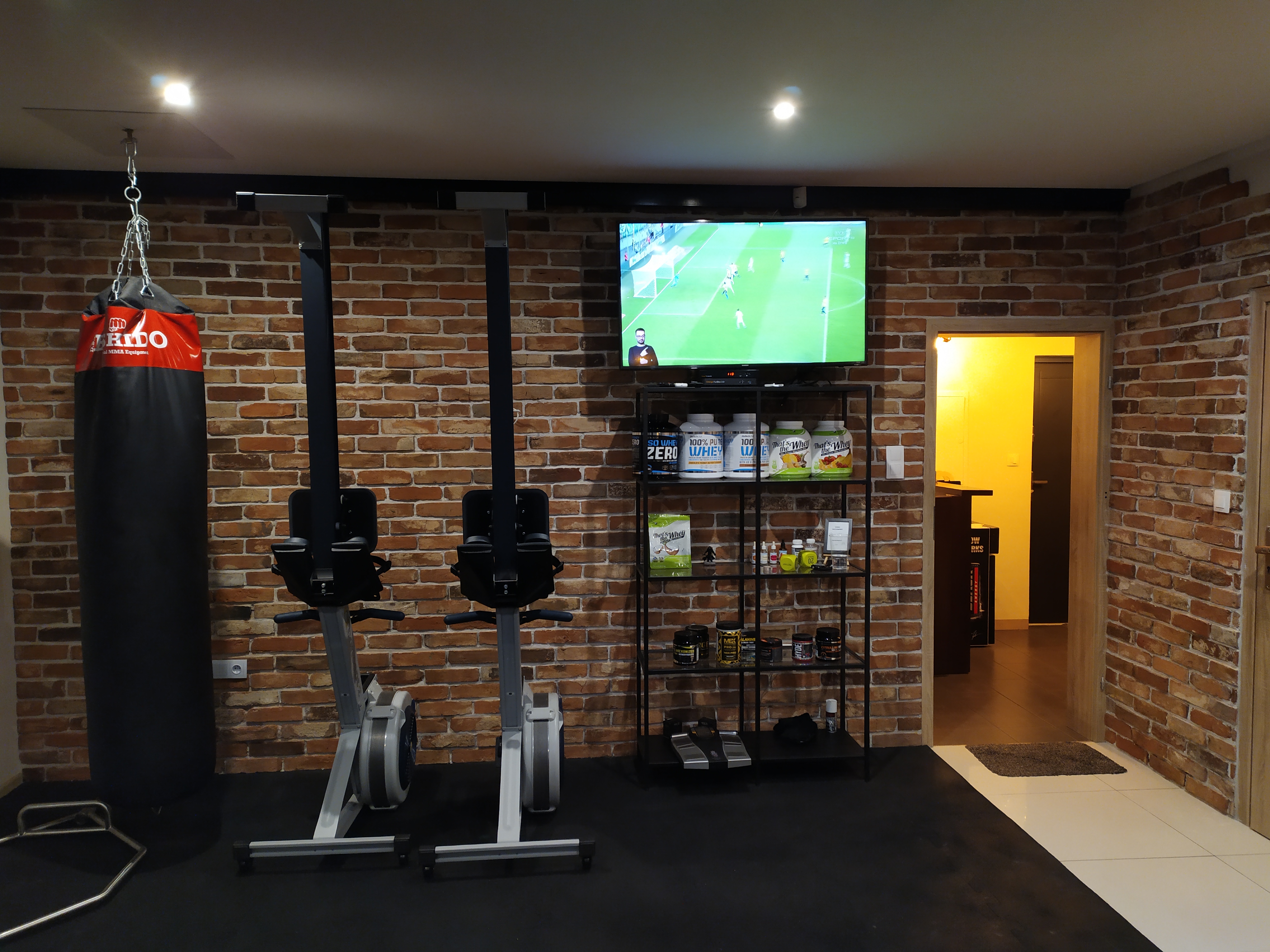 Domowa siłownia z dwoma ergometrami wioślarskimi, workiem bokserskim, półką z suplementami i telewizorem z meczem piłki nożnej, ceglana ściana w tle.