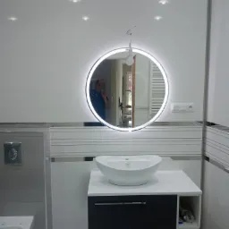 Nowoczesna łazienka z okrągłym lustrem LED, białą ceramiką i szarymi płytkami, widoczne odbicie fotografa w lustrze.