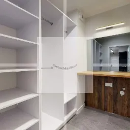 Biała, pusta garderoba z półkami i drążkami, widok na łazienkę z blatem z drewna i grzejnikiem drabinkowym w tle.