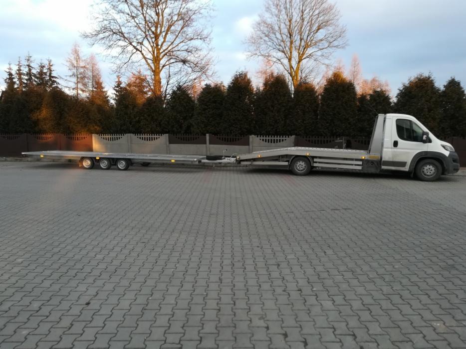 Długa laweta doczepiona do białego busa na kostce brukowej, z widocznym ogrodzeniem i drzewami w tle.