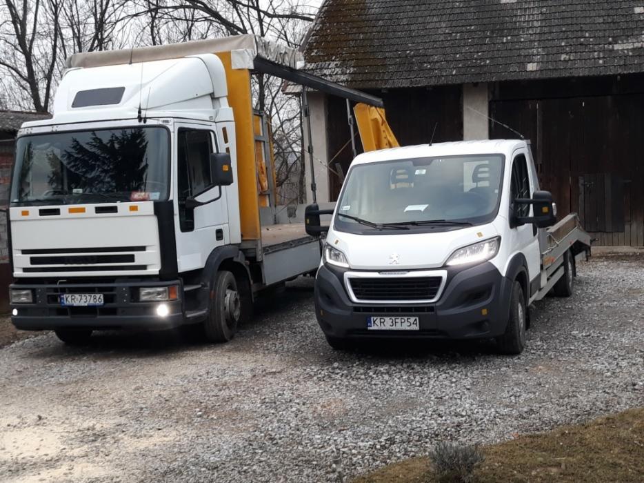 Dwa białe pojazdy: ciężarówka z żółtą plandeką i żurawiem oraz laweta na żwirowym podłożu przed starym budynkiem.