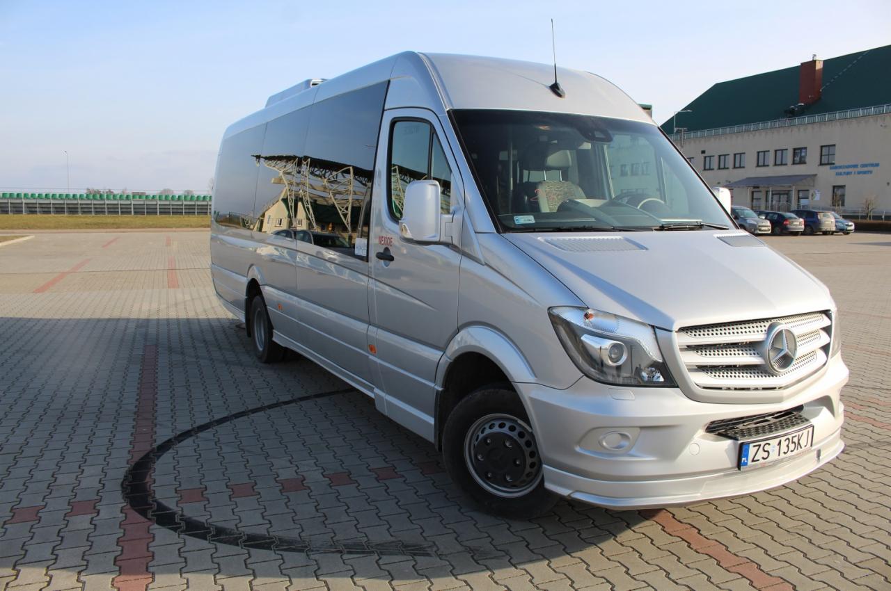 Srebrny bus Mercedes-Benz Sprinter z ciemnymi szybami na parkingu z kostki brukowej, widoczne ślady opon na kostce, w tle budynek z napisem 'Ośrodek Sportu i Rekreacji'.