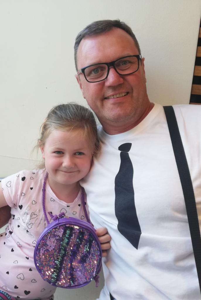 Uśmiechnięty mężczyzna w okularach i koszulce z krawatem pozuje z uśmiechniętą dziewczynką z brokatową torebką na tle jasnej ściany.