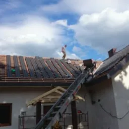 Układanie czarnej dachówki na drewnianej konstrukcji dachu domu jednorodzinnego. Prace na wysokości, widoczne drabiny i pracownicy. Częściowo pokryty dach.