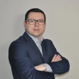 Portret adwokata w okularach, ubrany w granatową marynarkę i koszulę w kratę, z założonymi rękami na tle szarej ściany.