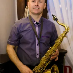 Portret mężczyzny z saksofonem, ubrany w fioletową koszulę z napisem MAX-MUSIC, na tle zasłon w odcieniach brązu i bieli.