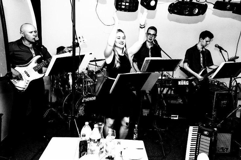 Czarno-białe zdjęcie zespołu muzycznego podczas występu: wokalistka z podniesionymi rękami, gitarzysta, basista i klawiszowiec, instrumenty Korg i Sonor, stolik z napojami w tle.