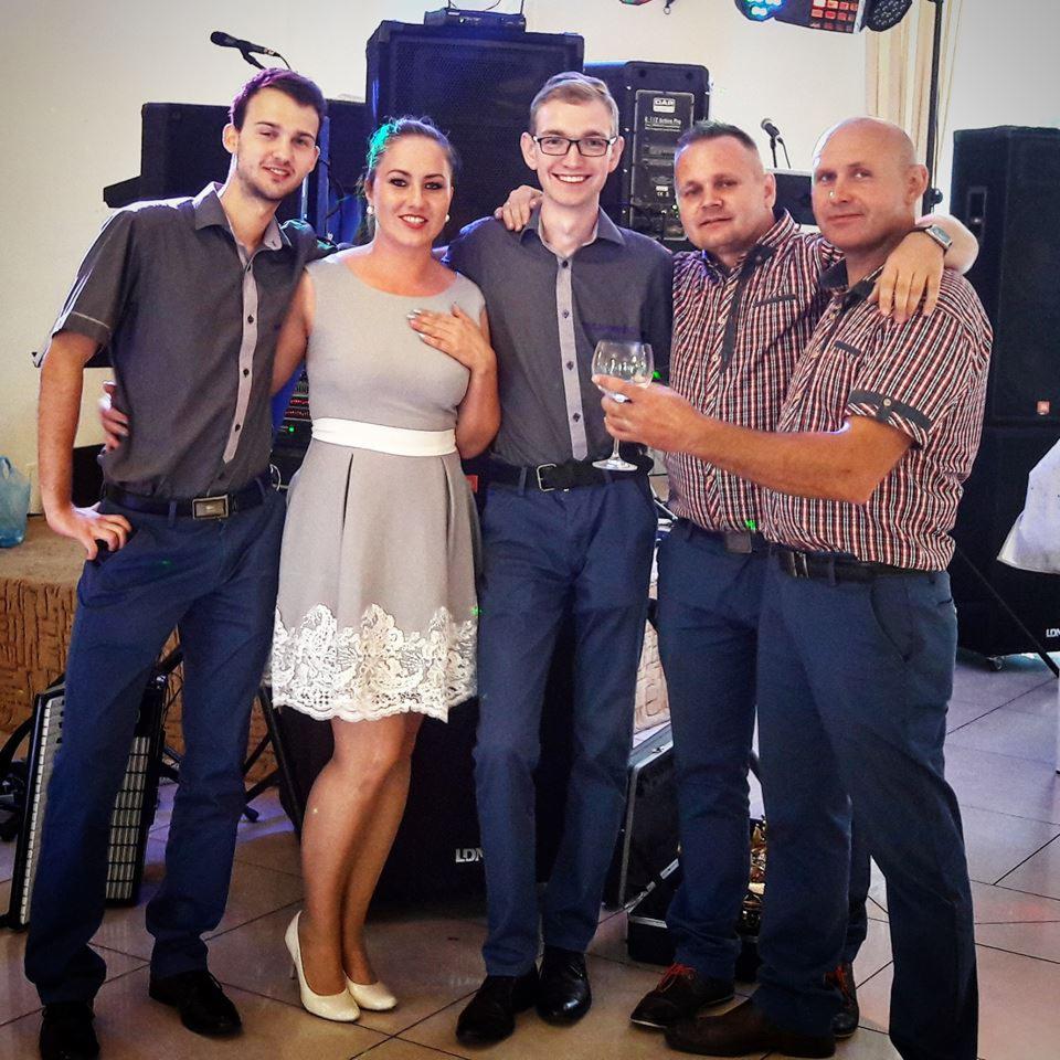 Pięcioosobowa grupa wokalno-instrumentalna w szaro-niebieskich strojach pozuje do zdjęcia na tle sprzętu nagłośnieniowego i oświetlenia scenicznego.