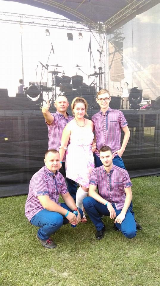 Pięcioosobowa grupa w dopasowanych koszulach w kratę i niebieskich spodniach pozuje na trawie przed sceną z zestawem perkusyjnym w tle.