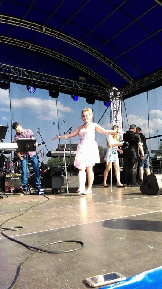 Wokalistka w białej sukience na scenie, z podpartą nogą kulami, obok muzycy i instrumenty, widoczny przewód na podłodze i telefon komórkowy.